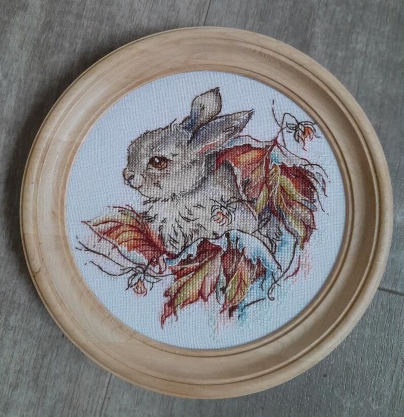 Peut inclure: Une broderie au point de croix encadr&eacute;e repr&eacute;sentant un petit lapin blanc assis parmi des feuilles d'automne color&eacute;es. Le lapin a des yeux bruns et une queue touffue.
