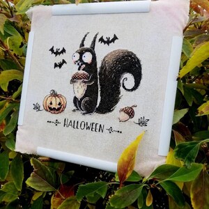 May include: Cross-stitch artwork featuring a black squirrel holding an acorn, with a jack-o'-lantern and bats. The word "HALLOWEEN" is stitched below the squirrel. The artwork is mounted on a white frame, set against a backdrop of green and yellow foliage.
