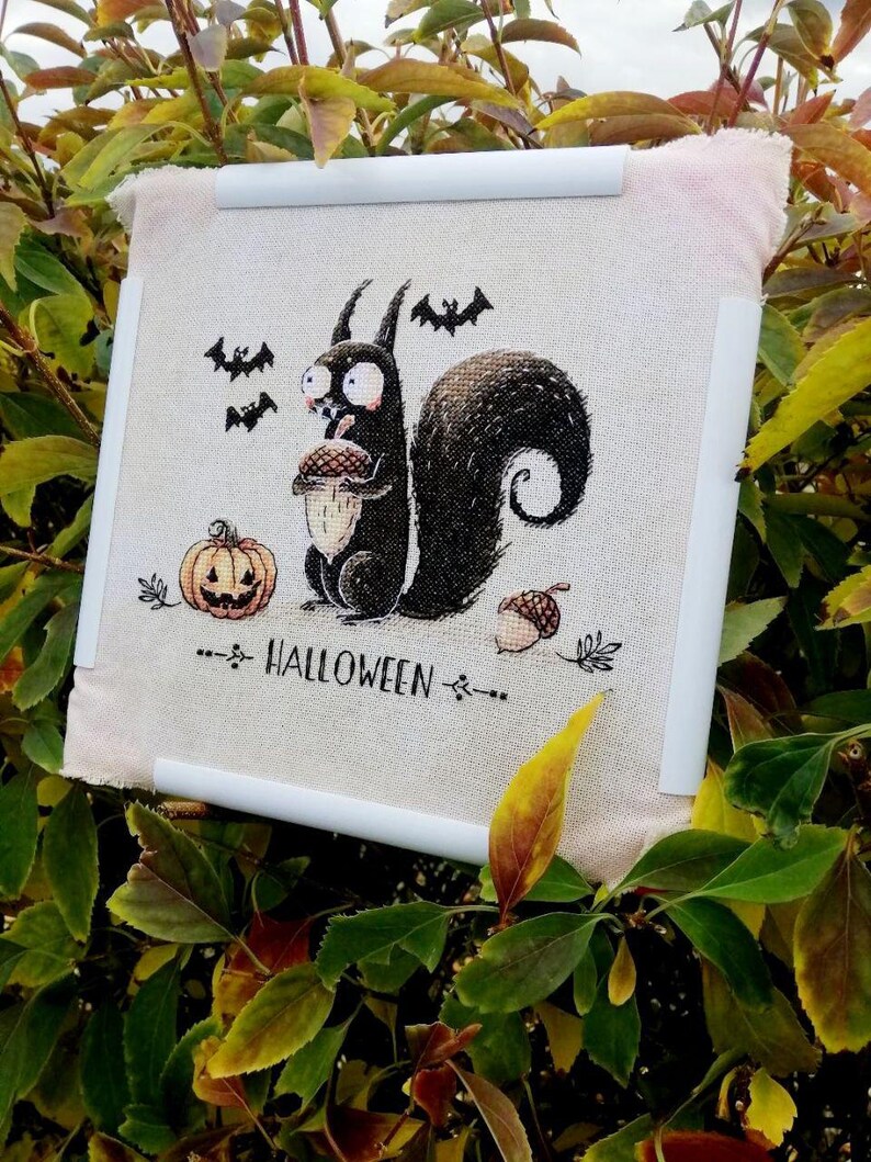 May include: Cross-stitch artwork featuring a black squirrel holding an acorn, with a jack-o'-lantern and bats. The word "HALLOWEEN" is stitched below the squirrel. The artwork is mounted on a white frame, set against a backdrop of green and yellow foliage.