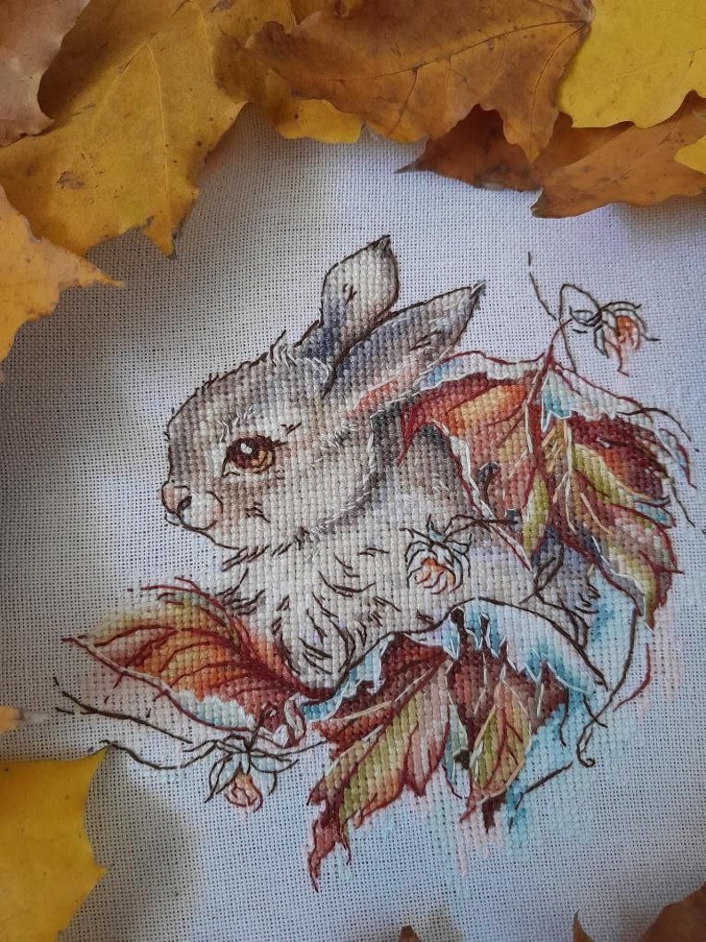 Peut inclure: Une broderie au point de croix repr&eacute;sentant un lapin gris assis parmi des feuilles d'automne color&eacute;es. Le lapin a des yeux bruns et un nez blanc. Les feuilles sont de couleur orange, rouge et jaune.