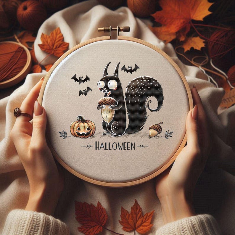 May include: A Halloween-themed cross-stitch embroidery hoop featuring a black squirrel holding an acorn, a jack-o'-lantern, and bats. The word "HALLOWEEN" is stitched below the squirrel. The embroidery is set in a wooden hoop, surrounded by autumn leaves.