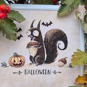 May include: Cross-stitch artwork featuring a cartoon squirrel holding an acorn, a jack-o'-lantern, and bats. The word "HALLOWEEN" is stitched below the image. The artwork is framed in a white frame and surrounded by green leaves and red berries.