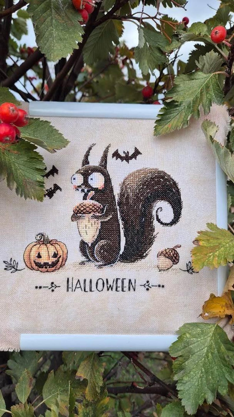 May include: Cross-stitch artwork featuring a cartoon squirrel holding an acorn, a jack-o'-lantern, and bats. The word "HALLOWEEN" is stitched below the image. The artwork is framed in a white frame and surrounded by green leaves and red berries.