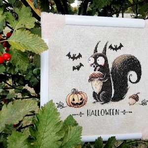 May include: A framed Halloween-themed cross-stitch featuring a cartoon squirrel holding an acorn, with a jack-o'-lantern, bats, and the word "HALLOWEEN". The artwork is framed in white with a light beige fabric background.