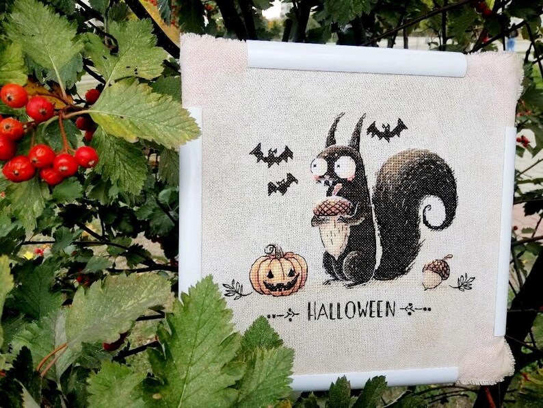 May include: A framed Halloween-themed cross-stitch featuring a cartoon squirrel holding an acorn, with a jack-o'-lantern, bats, and the word "HALLOWEEN". The artwork is framed in white with a light beige fabric background.