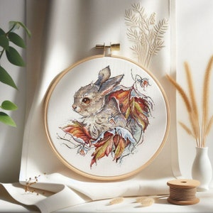 Peut inclure: Un cerceau de broderie au point de croix repr&eacute;sentant un lapin brun entour&eacute; de feuilles d'automne color&eacute;es. Le lapin regarde vers la gauche et a une queue blanche et moelleuse.