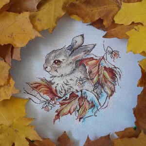 Peut inclure: Une broderie au point de croix repr&eacute;sentant un lapin blanc entour&eacute; de feuilles d'automne. Le lapin regarde vers la gauche et a des yeux bruns. Les feuilles sont dans des tons d'orange, de jaune et de rouge.
