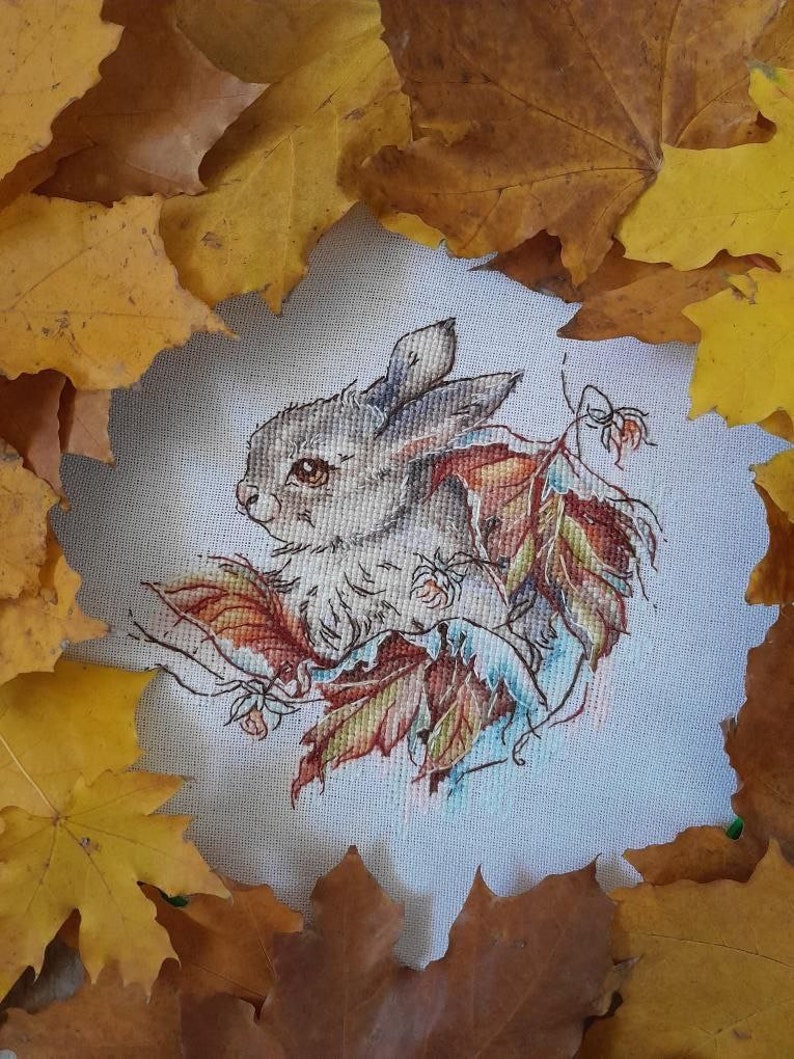 Peut inclure: Une broderie au point de croix repr&eacute;sentant un lapin blanc entour&eacute; de feuilles d'automne. Le lapin regarde vers la gauche et a des yeux bruns. Les feuilles sont dans des tons d'orange, de jaune et de rouge.