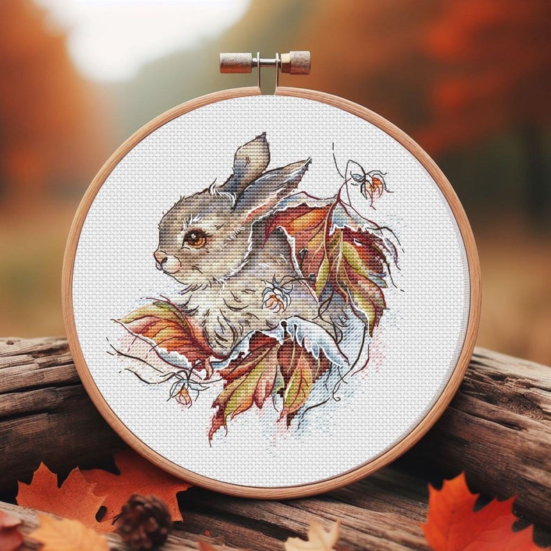 Peut inclure: Un cerceau de broderie en point de croix avec un fond blanc repr&eacute;sentant un lapin brun entour&eacute; de feuilles d'automne color&eacute;es. Le lapin regarde vers la gauche et a un nez brun et des yeux bruns.