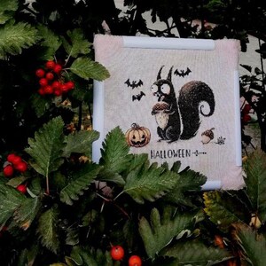 May include: A Halloween-themed cross-stitch design featuring a black squirrel holding an acorn, with bats and a jack-o'-lantern. The word "HALLOWEEN" is stitched below the squirrel. The design is framed in a white and pink border, set against a backdrop of green leaves and red berries.
