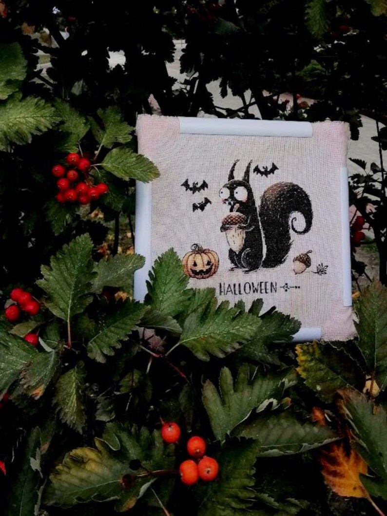 May include: A Halloween-themed cross-stitch design featuring a black squirrel holding an acorn, with bats and a jack-o'-lantern. The word "HALLOWEEN" is stitched below the squirrel. The design is framed in a white and pink border, set against a backdrop of green leaves and red berries.
