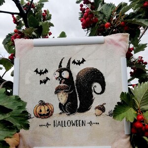 May include: A cross-stitch piece featuring a black squirrel holding an acorn, a jack-o'-lantern, and bats. The word "HALLOWEEN" is stitched below the squirrel. The piece is framed with white bars and surrounded by red berries and green leaves.