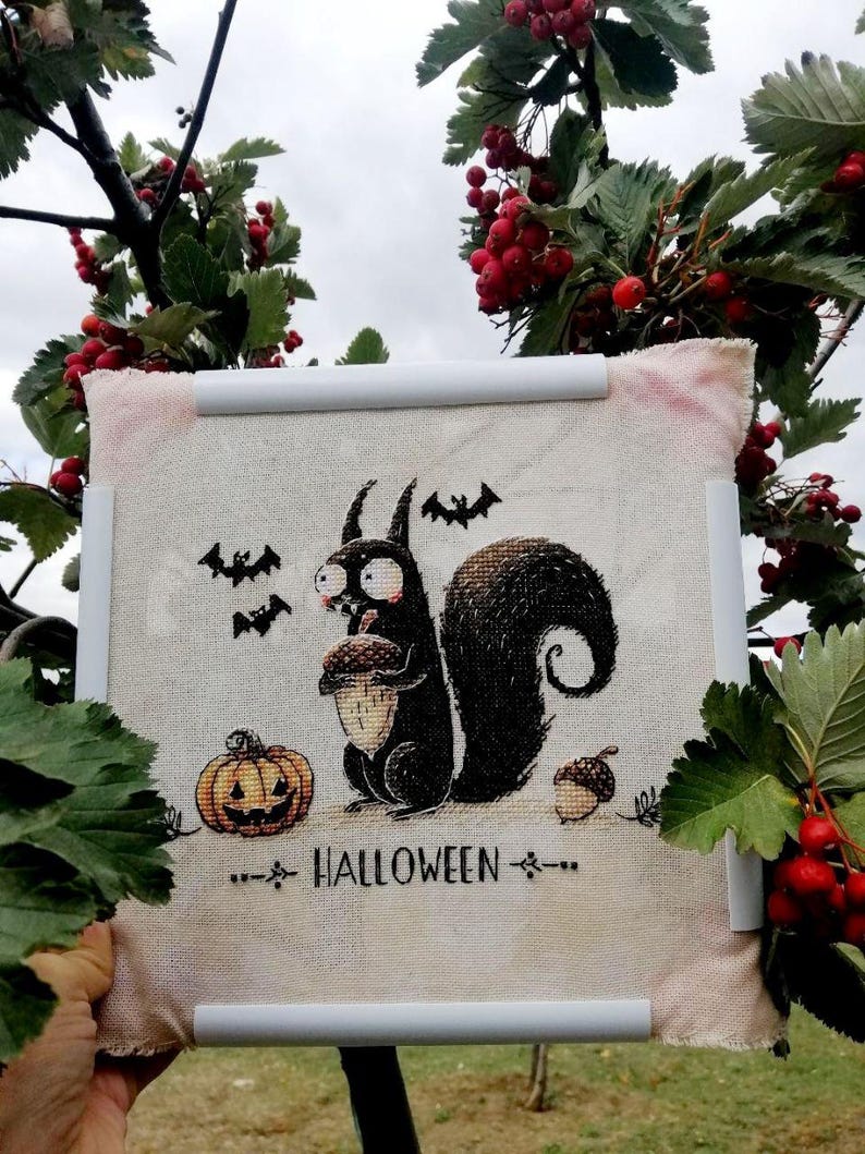 May include: A cross-stitch piece featuring a black squirrel holding an acorn, a jack-o'-lantern, and bats. The word "HALLOWEEN" is stitched below the squirrel. The piece is framed with white bars and surrounded by red berries and green leaves.