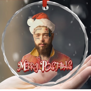 May include: A clear, round Christmas ornament featuring a portrait of a man wearing a Santa hat and a red jacket. The ornament has the text "MERRY POSTMAS" in red and white. A red ribbon is attached for hanging.