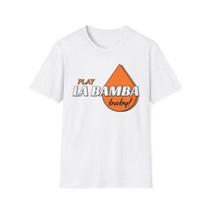 Könnte beinhalten: Weißes T-Shirt mit dem Text "PLAY LA BAMBA baby!" in Schwarz und Orange. Das Design zeigt eine stilisierte orangefarbene Tropfenform. Das Shirt hat kurze Ärmel und einen Rundhalsausschnitt.