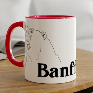 May include: White ceramic mug with a red rim. The mug features a line drawing of a bear with its mouth open, and the word "Banff" in black lettering.
