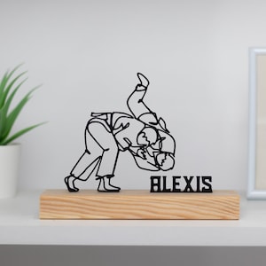 Op de afbeelding: Een zwart draadbeeld van twee mensen in een worstelpose op een houten basis met de naam "ALEXIS" erop gedrukt.