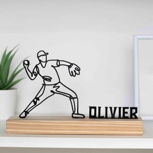 Op de afbeelding: Een zwarte draadsculptuur van een honkbalspeler in een worphouding, gemonteerd op een licht houten voetstuk. De naam "OLIVIER" wordt in zwarte blokletters naast de sculptuur weergegeven. Een potplant en een ingelijste foto zijn op de achtergrond.