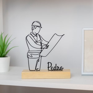 Puede incluir: Una escultura de alambre negro de una persona con un casco y sosteniendo un plano. La escultura está sobre una base de madera con el nombre "Pedro" escrito en ella.