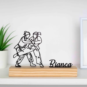 Op de afbeelding: Een zwarte metalen sculptuur van twee personen in een Judo-houding, gemonteerd op een rechthoekige houten basis. De naam "Bianca" wordt in cursief schrift naast de sculptuur weergegeven. De sculptuur staat tegen een witte achtergrond.