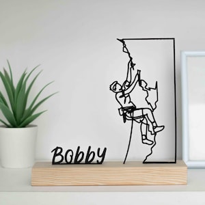 May include: Black metal sculpture depicting a climber on a rock face, set on a rectangular wooden base. The name "Bobby" is written in black cursive on the base. A potted green plant is in the background.