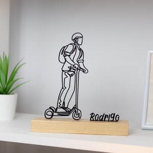 Könnte beinhalten: Schwarze Drahtskulptur einer Person, die einen E-Scooter mit Rucksack fährt. Die Skulptur steht auf einem Holzsockel mit dem Namen "Rodrigo" darauf.