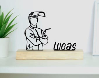 May include: A black metal sculpture of a welder with arms crossed, wearing a welding helmet, mounted on a rectangular wooden base. The name "Lucas" is written in black script next to the sculpture. A potted plant is visible on the left.