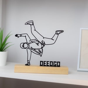 Könnte beinhalten: Schwarze Drahtskulptur einer Person, die Breakdance tanzt, auf einem Holzsockel mit dem Wort "DEEDIGO" darauf.