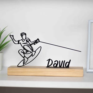 May include: Black metal wire sculpture of a person wakeboarding with the name "David" on a wooden base.