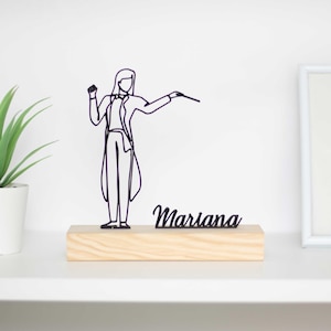 Könnte beinhalten: Eine schwarze Drahtskulptur einer Person, die ein Orchester dirigiert, steht auf einem Holzsockel mit dem Namen "Mariana" in schwarzen Lettern.