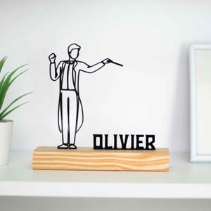 Może przedstawiać: Czarna rzeźba z drutu przedstawiająca dyrygenta z batutą, stojąca na drewnianej podstawie z napisem "OLIVIER" czarnymi literami.