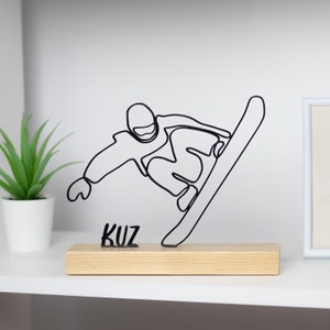 May include: Black wire sculpture of a snowboarder on a wooden base. The sculpture is titled "KUZ".