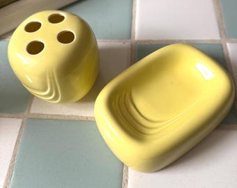 May include: A yellow ceramic toothbrush holder with four holes and a matching soap dish. The bathroom accessories are on a tiled surface with blue and white squares. The toothbrush holder is round and the soap dish is rectangular.