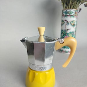 May include: A yellow and silver stovetop espresso maker with a yellow handle and top. The coffee maker has a classic design and is made of metal. A vase with flowers is in the background.