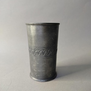 Puede incluir: Una taza cilíndrica de peltre con una banda decorativa de diseños curvos repetidos. La taza tiene una base ligeramente ensanchada y un borde enrollado. El metal tiene un acabado mate y un color gris.
