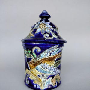 May include: A dark blue ceramic jar with a lid, decorated with a bird and floral motif. The jar is cylindrical, with a lid featuring a floral design. The bird is rendered in brown, blue, and gold tones, adding a touch of elegance.