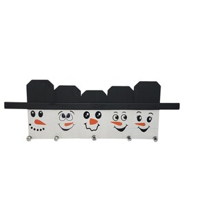 May include: A white wooden snowman-shaped key holder with five snowman faces. Each face has black eyes, a red nose, and a black mouth. The snowman faces are separated by black hat brims.