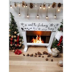 Puede incluir: Una repisa de chimenea navideña de madera blanca con el texto "...and the stockings were hung" y siluetas de copos de nieve y renos. Un pequeño árbol de Navidad está a cada lado de la repisa. La chimenea tiene un fuego encendido en su interior.
