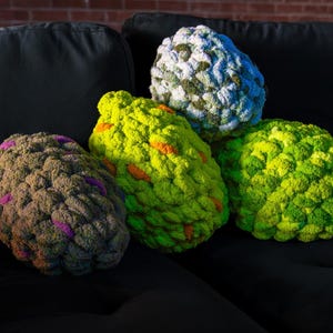 Pode incluir: Quatro almofadas de malha grossa em tons de verde, amarelo, azul e castanho. As almofadas têm um aspeto texturizado e irregular. Algumas almofadas têm detalhes em laranja e roxo. As almofadas estão dispostas numa superfície preta.