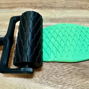 May include: A black rolling pin with a scale pattern is rolling over a green piece of clay, creating a scale pattern on the clay.