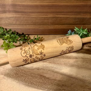 May include: A wooden rolling pin with engraved holiday-themed designs, including a snowman, reindeer, and snowflakes. The rolling pin is light brown and rests on a wooden surface, suggesting a kitchen or baking theme.