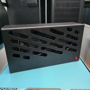 May include: A black rectangular metal enclosure with a series of cut-out shapes and a red power button. The enclosure sits on a light blue surface with the text "320 x 325 mm".