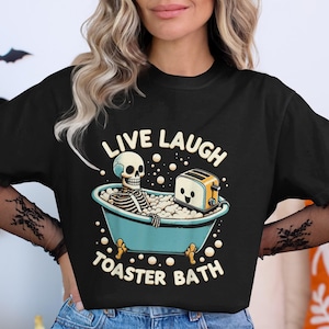 May include: Black t-shirt with a humorous graphic. The design features a skeleton in a blue bathtub with a smiling toaster, surrounded by bubbles. Text reads "Live Laugh Toaster Bath".