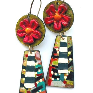 May include: Artistic dangle earrings featuring a red flower on a green circular base. Below, a colorful, abstract design with black, white, and teal stripes and dots. The earrings have gold-colored ear wires.
