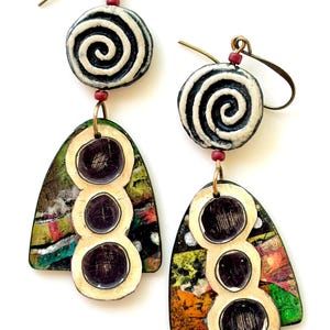 May include: Artistic dangle earrings with a spiral design and colorful geometric shapes. The earrings feature a black and white spiral at the top, connected to a colorful, abstract design with three dark circles. The earrings are suspended from gold-colored hooks.