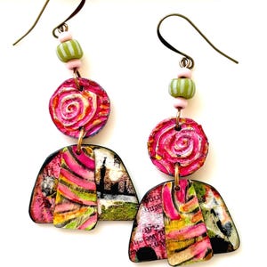 May include: A pair of handmade earrings with a unique, artistic design. The earrings feature a combination of pink, green, and yellow colors. Each earring has a circular top with a spiral design, connected to a fan-shaped bottom with abstract patterns and text.