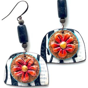 May include: A pair of handcrafted earrings featuring a black and white striped design. Each earring has a copper-colored circle with a red flower and a yellow center. The earrings have dark blue beads and metal hooks.