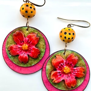 May include: A pair of handmade earrings featuring a red flower design. The earrings have a layered construction with a pink outer circle, a green inner circle, and a red flower. The top bead is yellow with black polka dots. The earrings hang from gold-colored hooks.