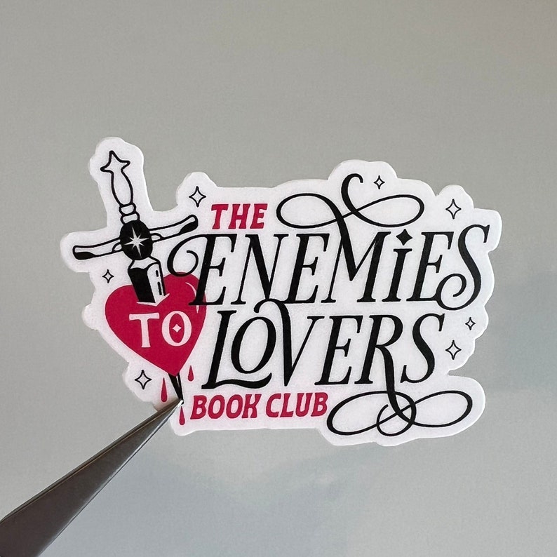 Peut inclure: Autocollant blanc avec du texte noir qui dit "The Enemies to Lovers Book Club" avec une fioriture stylis&eacute;e. Un c&oelig;ur rouge avec une dague noire qui le perce se trouve au centre de l'autocollant.