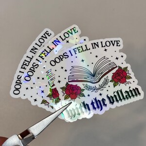 Puede incluir: Una pegatina hologr&aacute;fica con el texto "Oops I fell in love with the villain" y una ilustraci&oacute;n de un libro, rosas y estrellas.